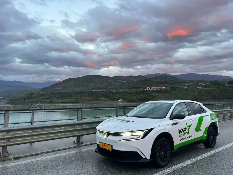 Electric taxi Elbasan — white and green Honda electric car on a bridge over a lake at sunset